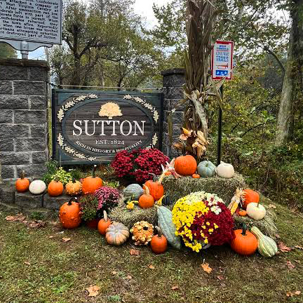Sutton, West Virginia town sign flanked on either side by cut stone pillars. in front of the sign sets a fall display of various pumpkins, gourds, and mums.